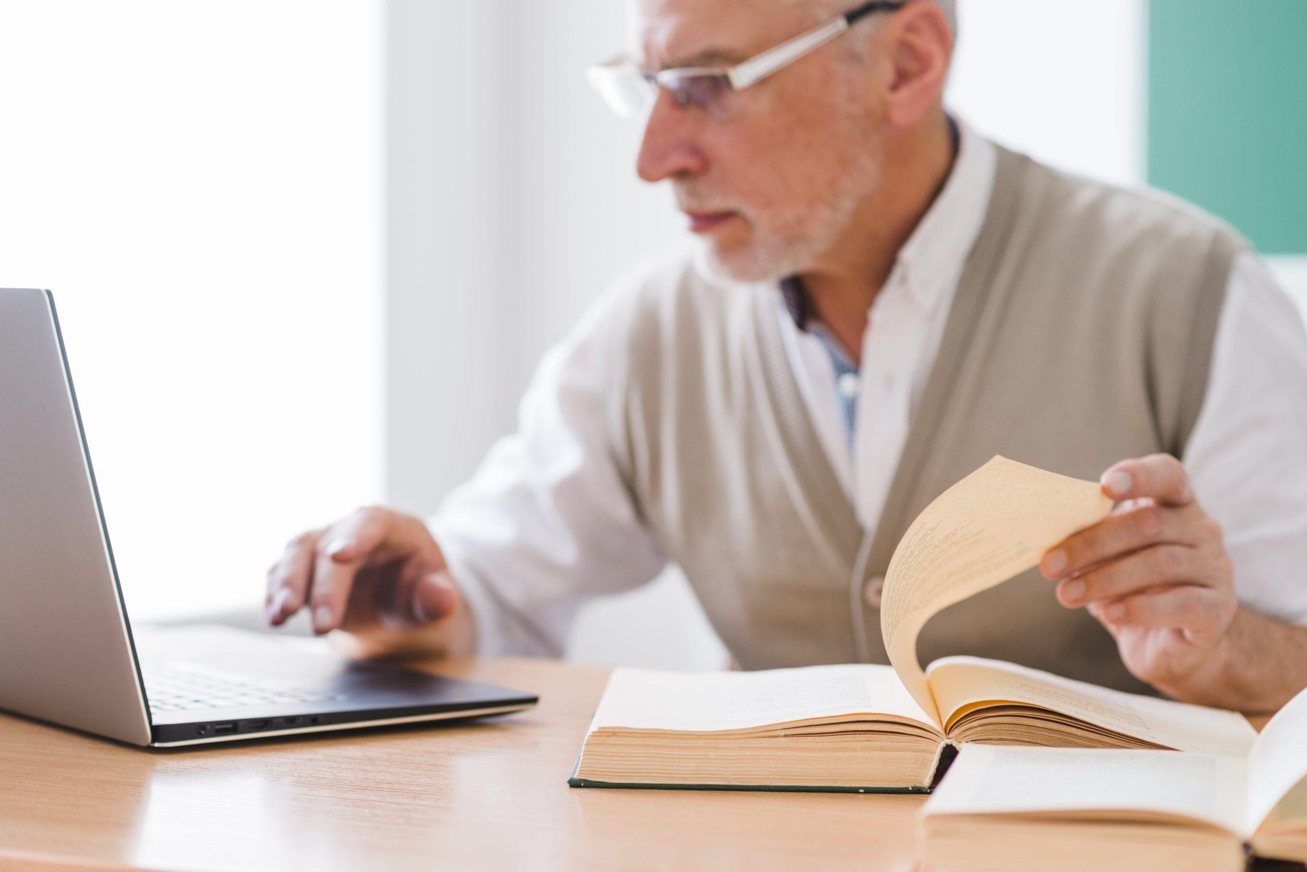 senior-professor-working-with-laptop-while-holding-page-of-book